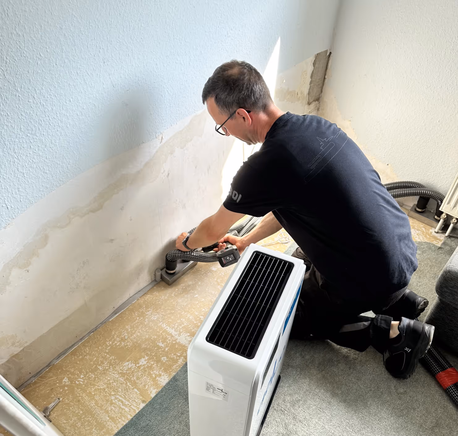 Ein Mann kniet am Boden und verbindet einen Schlauch mit einem tragbaren Klimagerät in einem Raum.
