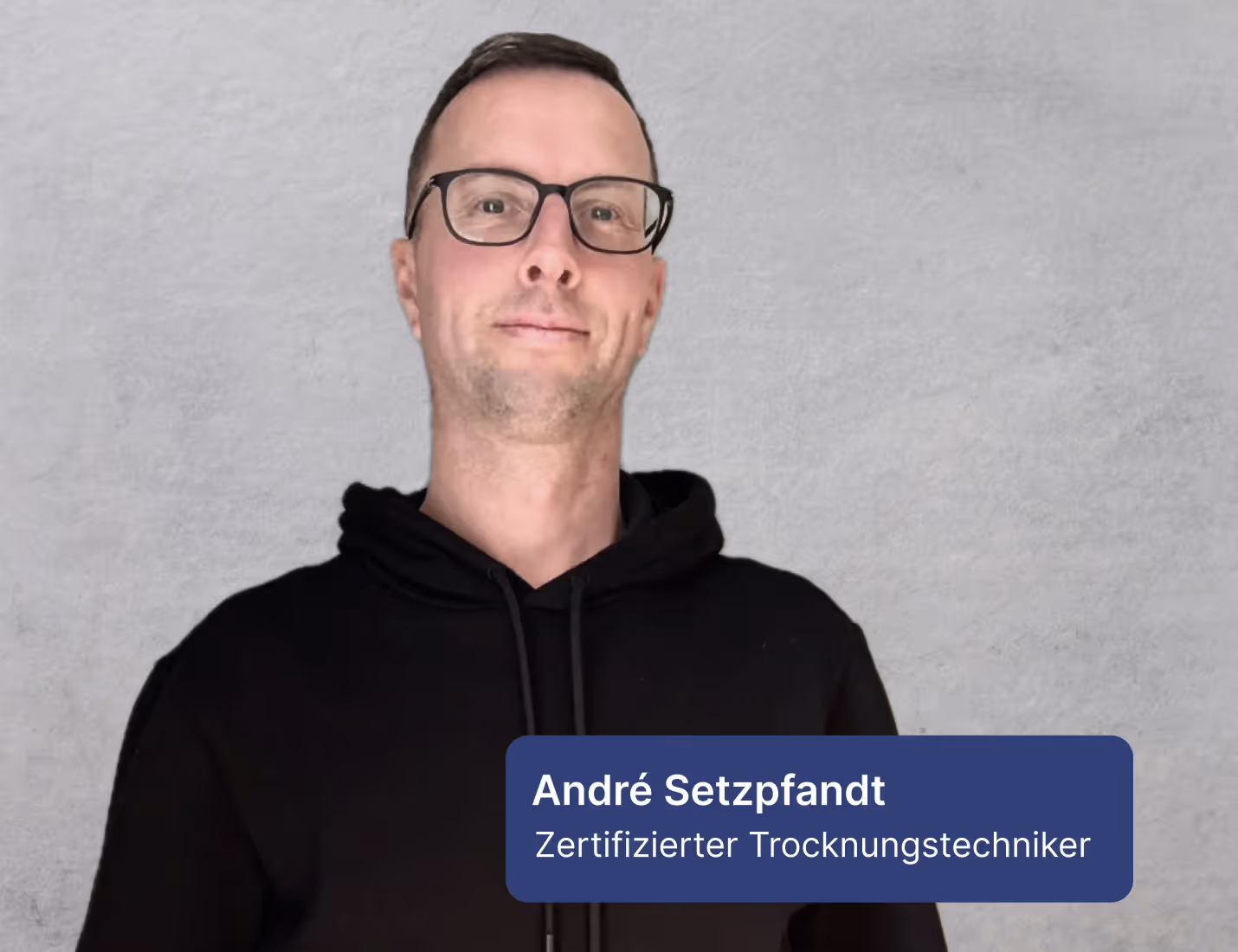 Porträt eines Mannes mit Brille und schwarzem Kapuzenpullover vor grauem Hintergrund.